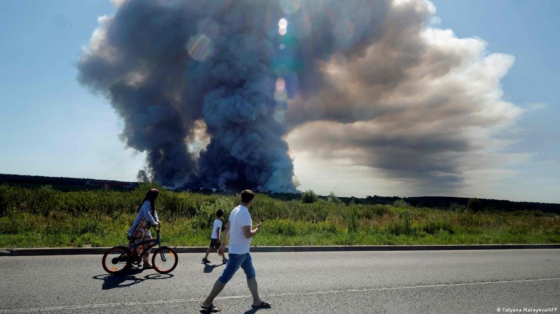 Personas pasan por una ruta, y en el horizonte se ve un iIncendio en el centro logístico de Wildberries tras un ataque con drones ucranianos. (18.07.2026).